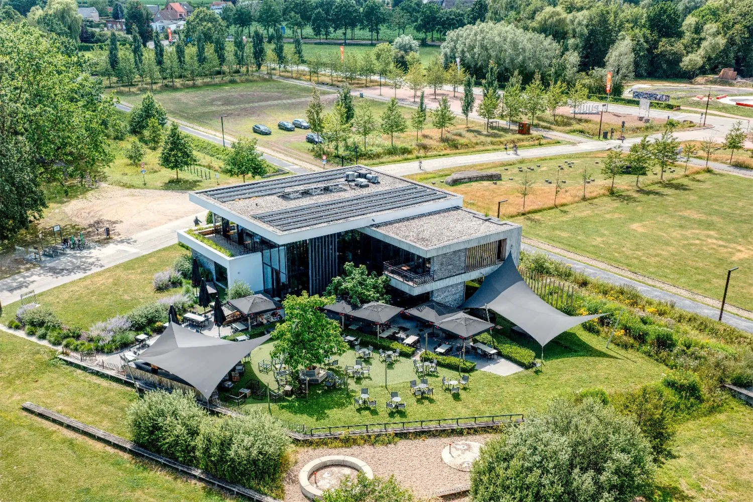 Luchtopname van een gebouw met twee platte daken, zonnepanelen, een glazen gevel en een terras met parasols en schaduwdoeken, gezien vanaf een inspectiedrone.